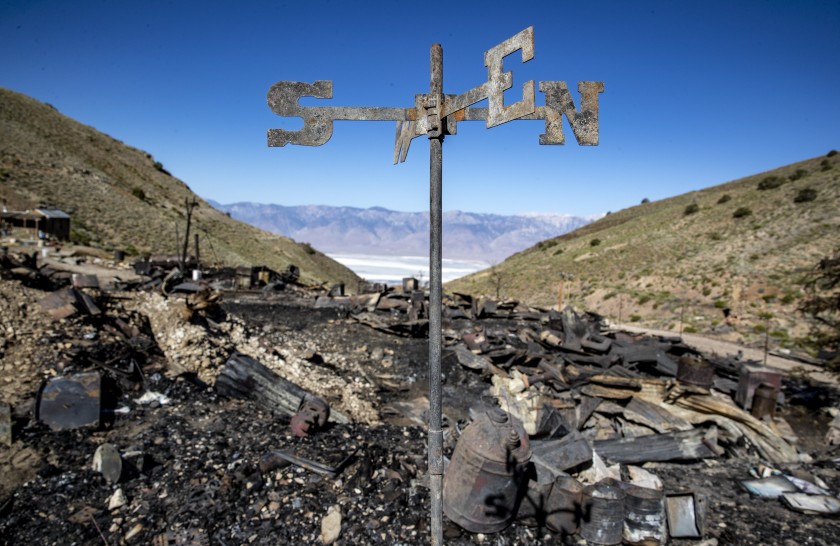 A weather vane blackened by fire stands among charred debris on a dry, brushy mountainside above a desert valley